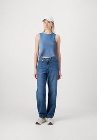 Blå ribbet crop-top, høy midje blå jeans og hvite joggesko. Modellen har på seg en hvit baseballcap. Minimalistisk bakgrunn.