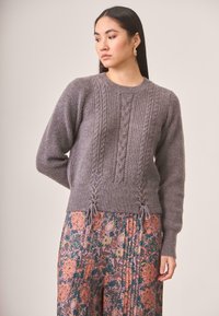 Suéter gris de punto con textura de canalé y detalle de trenzado, con cordones laterales, combinado con una falda con estampado floral.