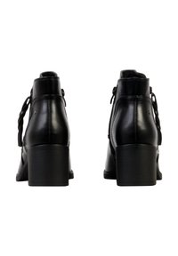 Paire de bottines en cuir noir à talons carrés vues de dos, montrant des détails de fermeture éclair et de pompon de chaque côté.