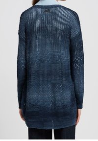 Kobieta ubrana w granatowy kardigan z otwartym splotem, nosząca jasnoniebieską koszulę z kołnierzykiem, pokazana od tyłu na jednolitym tle.