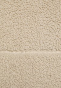 Beige fleece-tyg med en mjuk, strukturerad yta som har ett mönster av små, runda tofsar. Sömnad synlig längs mitten.