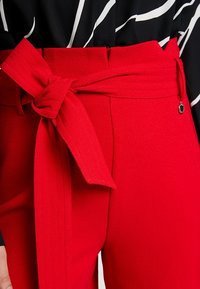 Vysoké červené kalhoty s širokým uvázaným páskem, nošené s topem s černobílým pruhovaným vzorem, zobrazení detailu v oblasti pasu.