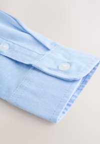 Poignet de chemise en tissu bleu clair avec un bouton blanc fermé, montrant les coutures détaillées et un tissage texturé sur une surface claire.