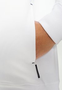 Ruka v boční kapse bílé košile s dlouhým rukávem, na lemu je viditelná etiketa "Scotch & Soda".