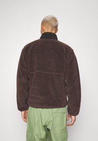 Brun fleecejacka med svart krage, som har ett texturerat, mjukt material, långa ärmar och en horisontell sömdetalj på baksidan.