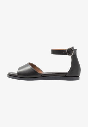 Svart lädersandal med öppen tå, rem runt vristen och rund spänne. Platt sula och slät yta; minimalistisk design.
