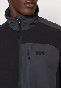 Veste zippée noire et grise avec col haut, dotée d'une poche poitrine zippée, d'un logo à l'avant et de panneaux en tissu texturé.