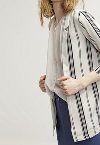 Světle barevný pruhovaný blazer s volným střihem, s černými vertikálními pruhy a 3/4 rukávy, navrstvený přes jednoduchý zapínací top.