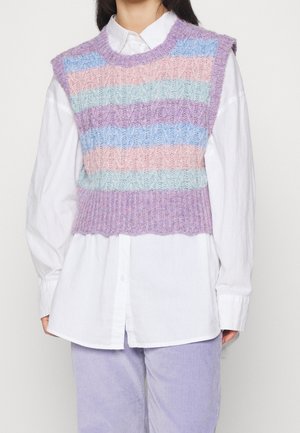 Person iført pastelfarvet stribet strikket vest over en hvid skjorte med knapper og lyse lilla fløjlsbukser, stående mod en ensfarvet baggrund.