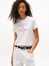 Hellgraues Baumwoll-T-Shirt mit kurzen Ärmeln und einem Grafikdruck, der "Tommy Jeans 1985" in Rot und Blau zeigt. Kombiniert mit weißen Jeans und einem schwarzen Gürtel.