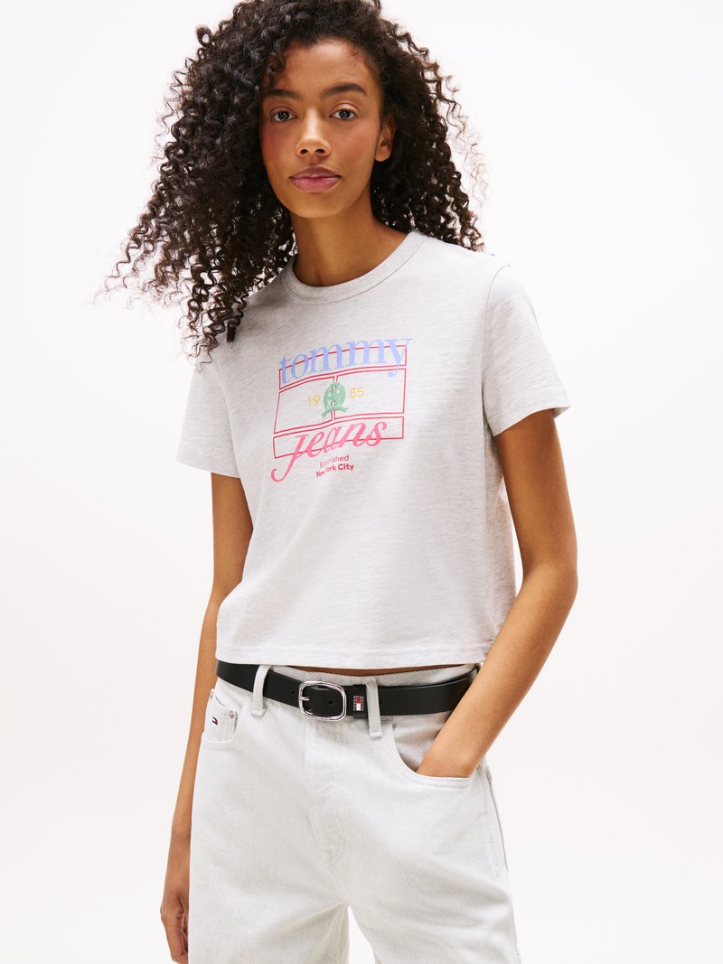 Jasnoszara bawełniana koszulka z krótkim rękawem, z graficznym nadrukiem "Tommy Jeans 1985" w kolorze czerwonym i niebieskim. Połączona z białymi dżinsami i czarnym paskiem.