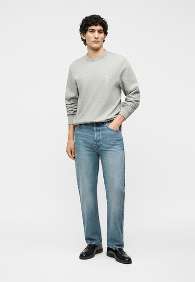 Νεαρός άνδρας που στέκεται με το χέρι στην τσέπη, φοράει ανοιχτό γκρι φούτερ, μπλε τζιν και μαύρα δερμάτινα παπούτσια σε απλό λευκό φόντο.