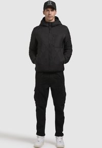 Schwarze Pufferjacke mit hexagonalem Muster, Reißverschluss und vorderer Tasche, kombiniert mit schwarzen Cargo-Hosen und weißen Sneakers.