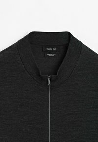Cardigan en laine mérinos gris foncé avec un col rond, fermeture éclair sur le devant et une texture en maille lisse. Étiquette visible à l'intérieur du col.
