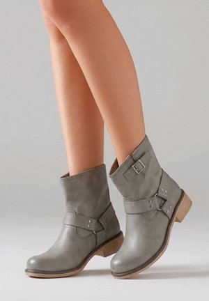 Botas de cuero gris hasta media pantorrilla con detalles de hebilla y correa, usadas en las piernas, con tacón bajo de madera y punta redondeada sobre un fondo liso.