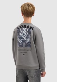 Persoon in een grijze sweatshirt met een stedelijke architectuurafbeelding en het woord "URBAN" op de achterkant, staand tegen een effen achtergrond.
