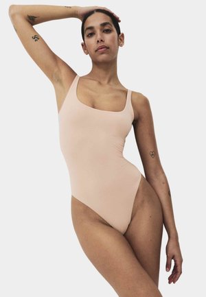 Kvinde med tatoveringer, der poserer i en lysebeige ærmeløs bodysuit mod en ensfarvet hvid baggrund.