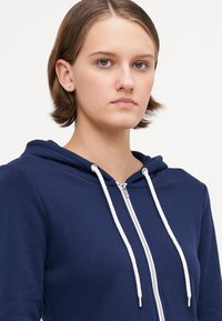 Marineblå hettegenser med glidelås, snor i hetten og hvite detaljer. Laget av myk stoff, med en jevn tekstur og en tettsittende passform.