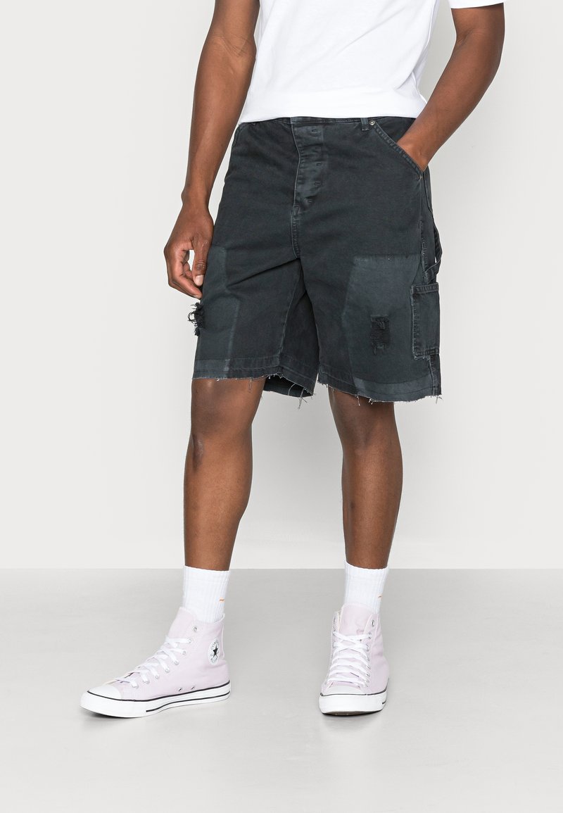Homme portant un short en denim noir usé, des chaussettes blanches montantes, des baskets montantes rose pâle et un t-shirt blanc, debout les mains dans les poches.