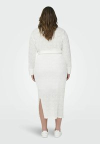 Witte kanten jurk met een getextureerd bloemmotief, lange mouwen en een midi lengte. Heeft een zijsplits en wordt gecombineerd met witte schoenen.