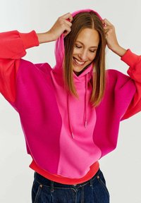 Kapsáčová mikina se stahovací kapucí s přechodem od růžových odstínů a oranžovým akcentem na lemu. Vyrobena z měkké tkaniny s oversize volným střihem.