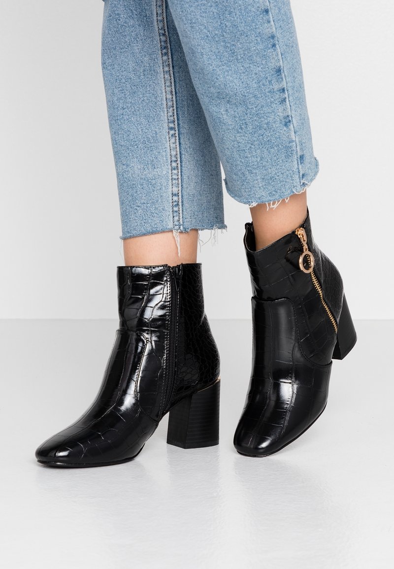 Svarta, lackskinnsanklar med krokodilmönster, med en sidodragkedja med ett cirkulärt handtag och en blockklack.