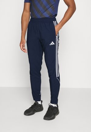 Homme portant un pantalon de survêtement Adidas bleu marine avec des bandes blanches et des baskets Adidas noires, debout devant un fond clair uni.