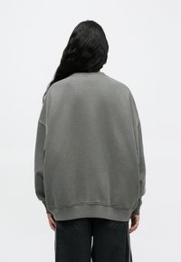 Άτομο με μακριά σκούρα μαλλιά που φοράει υπερμεγέθη γκρι φούτερ και μαύρο παντελόνι, γυρισμένο μπροστά σε απλό ανοιχτό φόντο.