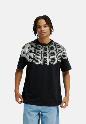Jeune personne aux tresses mi-longues portant un t-shirt noir avec un texte graphique blanc en gras et un jean bleu clair, debout devant un fond uni.