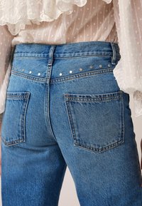 Jean en denim bleu taille haute avec des clous métalliques le long de la ceinture, associé à un chemisier blanc transparent avec des détails en dentelle florale brodée.