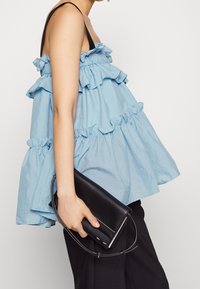 Top senza maniche blu chiaro con strati a volant, corpino aderente e orlo fluente, abbinato a una pochette nera tenuta in mano.