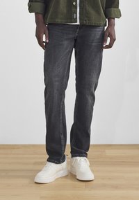 Black denim jeans with a slim fit, featuring a faded texture. Paired with white sneakers, standing on a wooden floor.