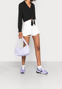 Schwarze Bluse mit langen Ärmeln und Knotendetail, weiße distressed Shorts, lavendelfarbene Hobo-Tasche und lavendelfarbene Sportschuhe mit schwarzen Akzenten.