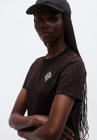 Brūns kokvilnas T-krekls ar dimanta formā darinātu baltu svītru izšuvumu uz krūtīm. Modelis valkā melnu cepuri un ar taisnām pītām matiem.