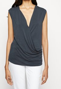 Dunkelgrau ärmelloses Top mit tiefem V-Ausschnitt und drapierter Vorderseite, aus weichem Stoff, kombiniert mit weißen Hosen.