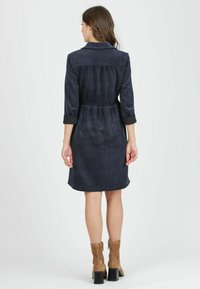 Dunkelblauses Corduroykleid mit Dreiviertelärmeln, tailliertem Bund und geschwungener Säume. Kombiniert mit hellbraunen Ankle Boots. Glatte Textur.