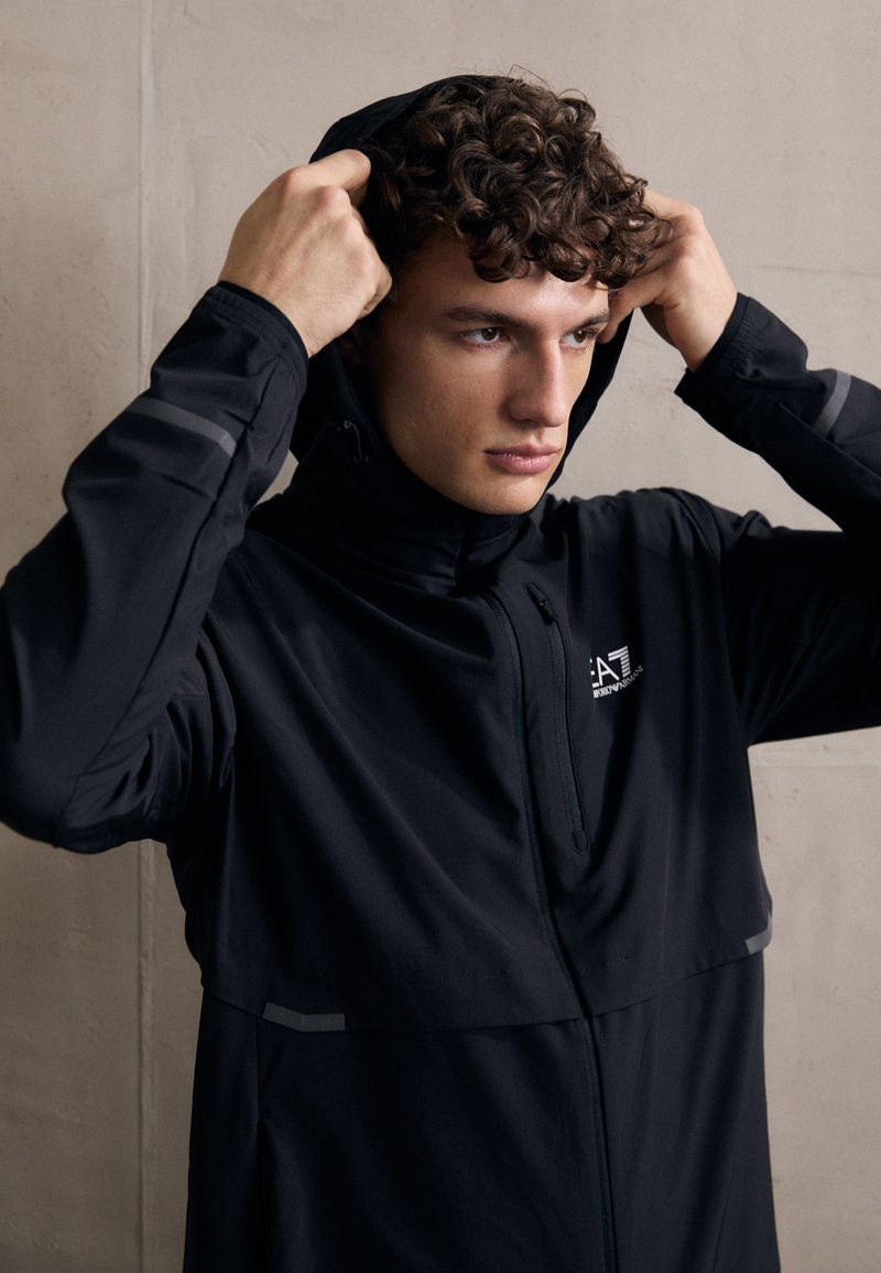 Μαύρο ελαφρύ μπουφάν με μπροστινό φερμουάρ και ψηλό γιακά. Διαθέτει ανακλαστικές ρίγες στα μανίκια και κουκούλα που έχει τραβηχτεί πάνω από το κεφάλι του μοντέλου.