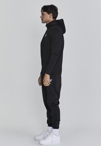 Zwarte capuchontrui en bijpassende joggers van gladde stof, met witte logo-accenten. Model draagt witte sneakers en crew-sokken.