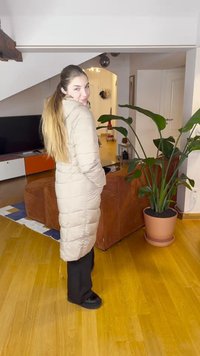 Geacă matlasată bej cu un design quiltat, lungime până la genunchi, buzunare laterale, asortată cu pantaloni și pantofi negri, într-un spațiu interior bine luminat.