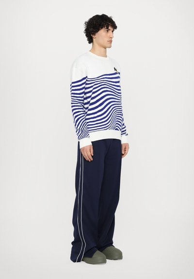 Blau-weiß gestreifter Strickpullover mit langen Ärmeln, kombiniert mit blauen Hosen und grünen Slippern. Weißer Kragen und gestreiftes Design.