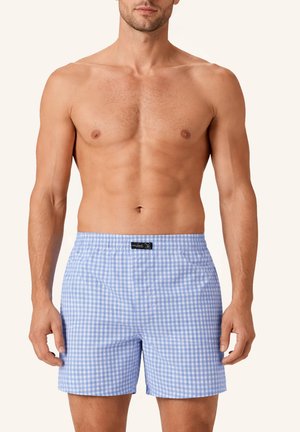 Muskulöser Mann trägt hellblaue und weiß karierte Boxershorts, steht vor einem schlichten beigen Hintergrund.