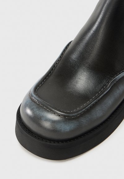 Μαύρο δερμάτινο μποτάκι στους αστραγάλους με λεία επιφάνεια, στρογγυλεμένη μύτη και παχύ λαστιχένιο πέλμα. Διαθέτει ορατή ρα φή κατά μήκος των άκρων.