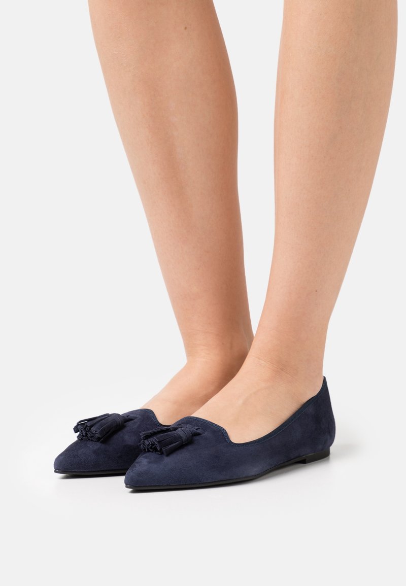 Chaussures plates en daim bleu marine avec un bout pointu, ornées d'un nœud décoratif. La semelle est plate et lisse, offrant un design minimaliste.