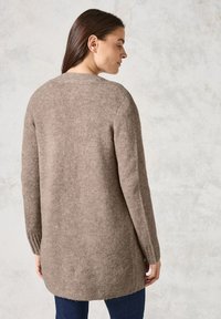 Cardigan long en maille tricotée douce de couleur brun clair, avec un col rond, des poignets côtelés et un ourlet légèrement arrondi.