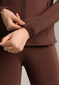 Smeđa sportska jakna s mekanim, postavljenim manžetama od flisa. Dizajn se odlikuje uskim krojem i glatkom teksturom, u kombinaciji s odgovarajućim smešim tajicama.