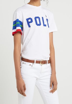 Personne portant une chemise blanche avec le texte bleu "POLO", des rayures multicolores aux manches, un pantalon blanc et une ceinture marron avec une boucle dorée.