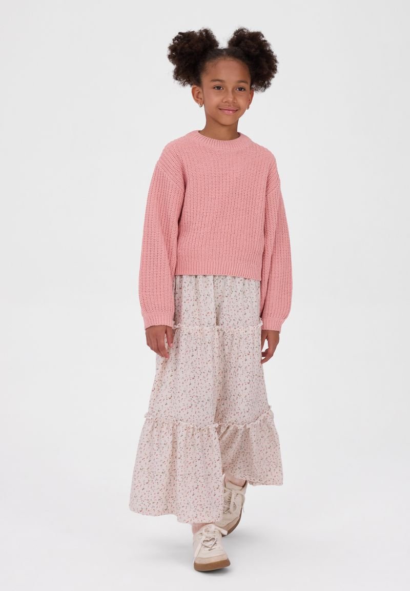 Jeune fille aux cheveux bouclés portant un pull rose, une longue jupe fleurie et des baskets blanches, debout sur un fond blanc uni.