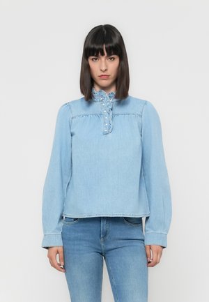 Blusa in denim azzurro chiaro con maniche lunghe, collo alto, dettagli con volant e bottoni decorativi sul davanti. Vestibilità casual.