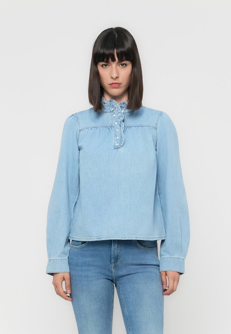 Light blue denim blouse with long sleeves, high neckline, ruffle detail, and decorative buttons along the front. Casual fit.