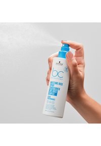 Flacon blanc avec buse de pulvérisation bleue, étiqueté "Après-shampoing en spray Moisture Kick", tenu dans une main, avec une fine brume se diffusant de la buse.
